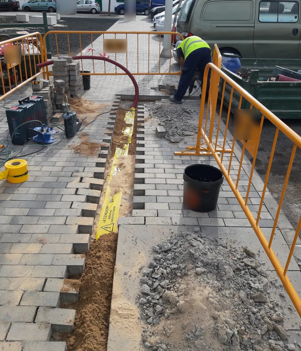Zanja de una canalización eléctrica abierta en una acera, con el vallado de la obra y la cinta de señalización sobre los tubos